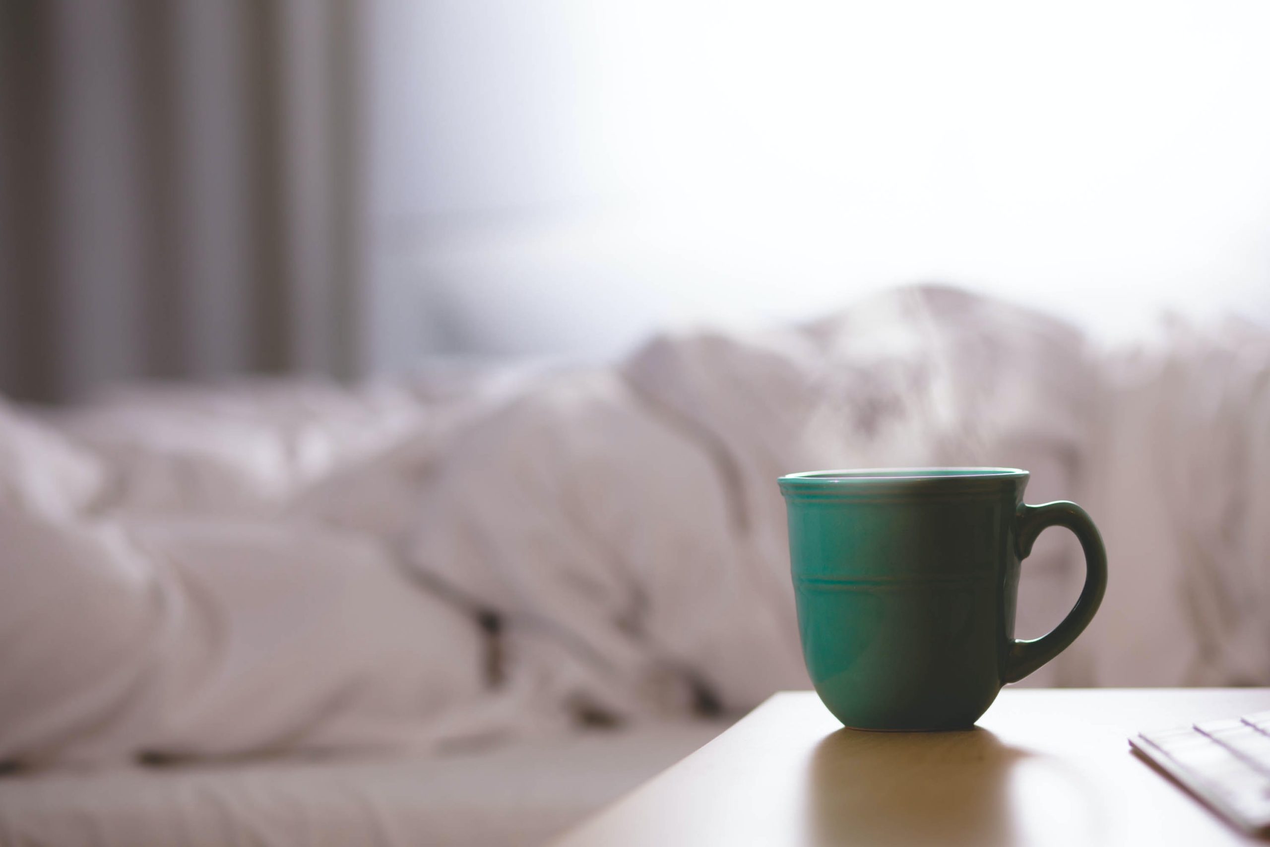 green mug with steam and white bed sheets in the background