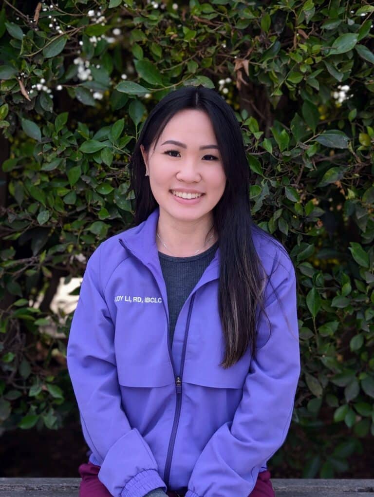 asian lactation consultant wearing purple FIGS scrub jacket sitting with green leaves in the background